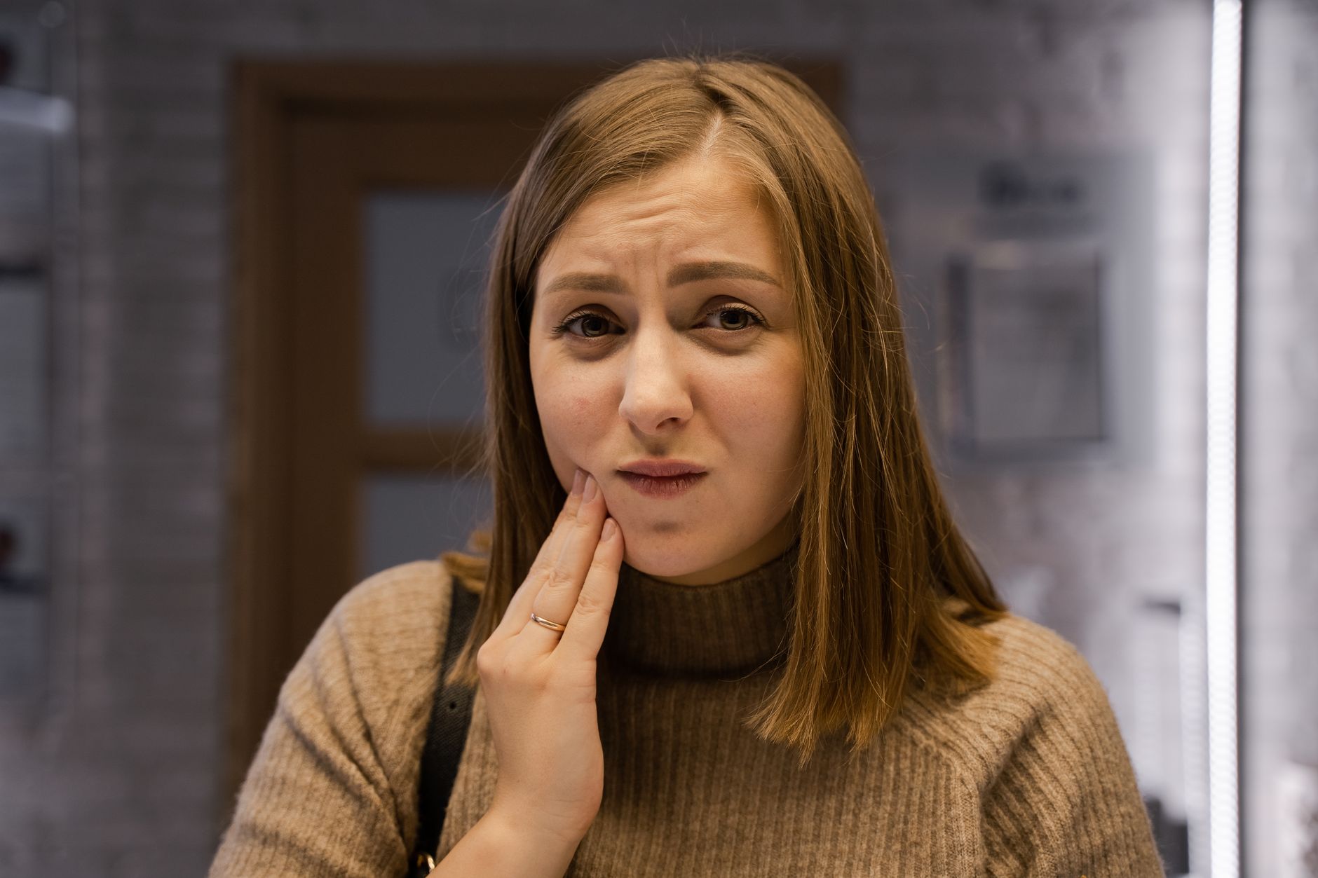 A woman in a brown sweater indoors holding her cheek, appearing to experience tooth discomfort.