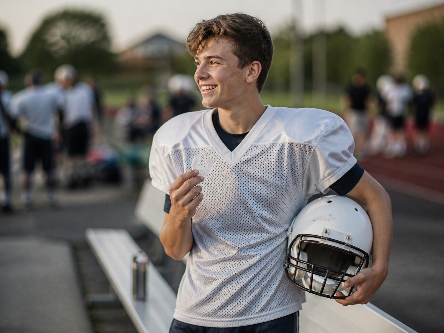 A smiling teenage athlete in football practice gear on the sideline, helmet under one arm, holding a sports mouthguard